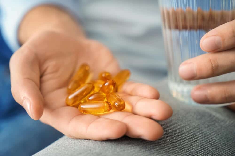 A person holds several yellow vitamin gel capsules—possibly a tab 4 supplement—in one hand and a glass of water in the other.