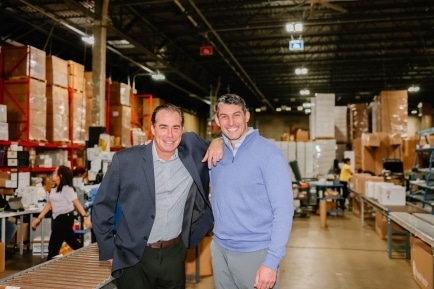 Two smiling men standing in a cold chain warehousing facility with shelves and packages in the background.