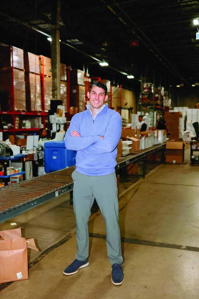 A man in a blue sweater and gray pants smiling and standing with crossed arms in a cold storage warehouse full of boxes and goods.