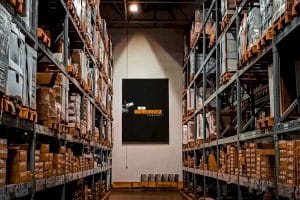 Warehouse interior with tall shelves stocked with boxes, a central aisle leading to a black sign promoting "The New Warehouse Podcast" featuring 3PL experts.