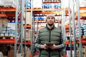 A man in a beanie and green vest holding a tablet, standing in a supply chain warehouse with tall shelves filled with boxes.