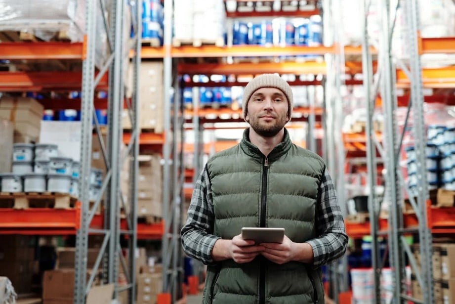 A man in a beanie and green vest holding a tablet, standing in a supply chain warehouse with tall shelves filled with boxes.