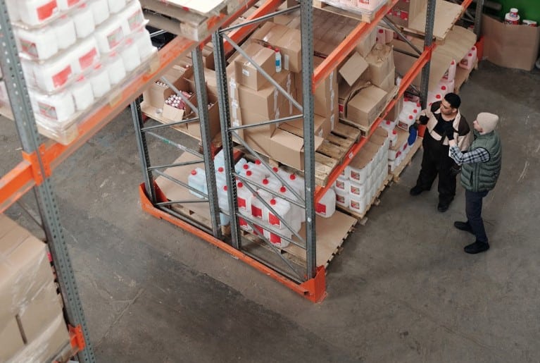 Two warehouse workers discussing inventory in a cold storage warehousing aisle with shelves stocked with various boxes and packages.