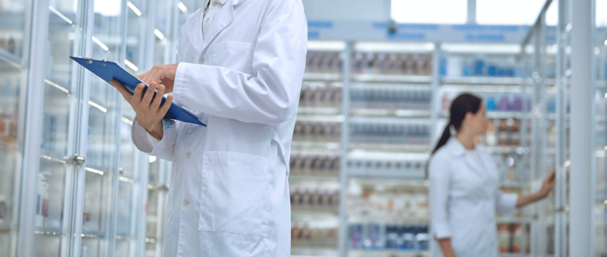 Two people in white lab coats work in a pharmacy; one holds a clipboard, while the other checks shelves stocked with medication, supplies, and items stored in pharmaceutical cold storage.
