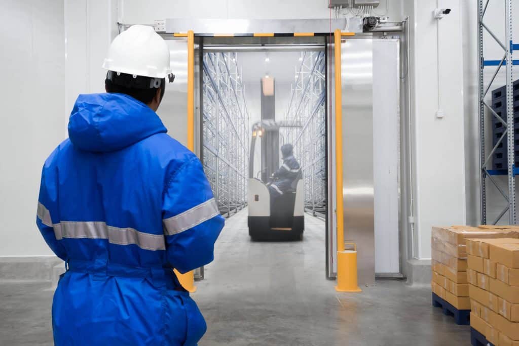 A worker in a blue jacket and white helmet watches a forklift transporting goods critical for cold chain food safety through an industrial warehouse doorway, with boxes stacked on the right.