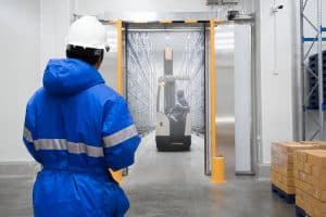 A worker in a blue jacket and white helmet watches a forklift transporting goods critical for cold chain food safety through an industrial warehouse doorway, with boxes stacked on the right.