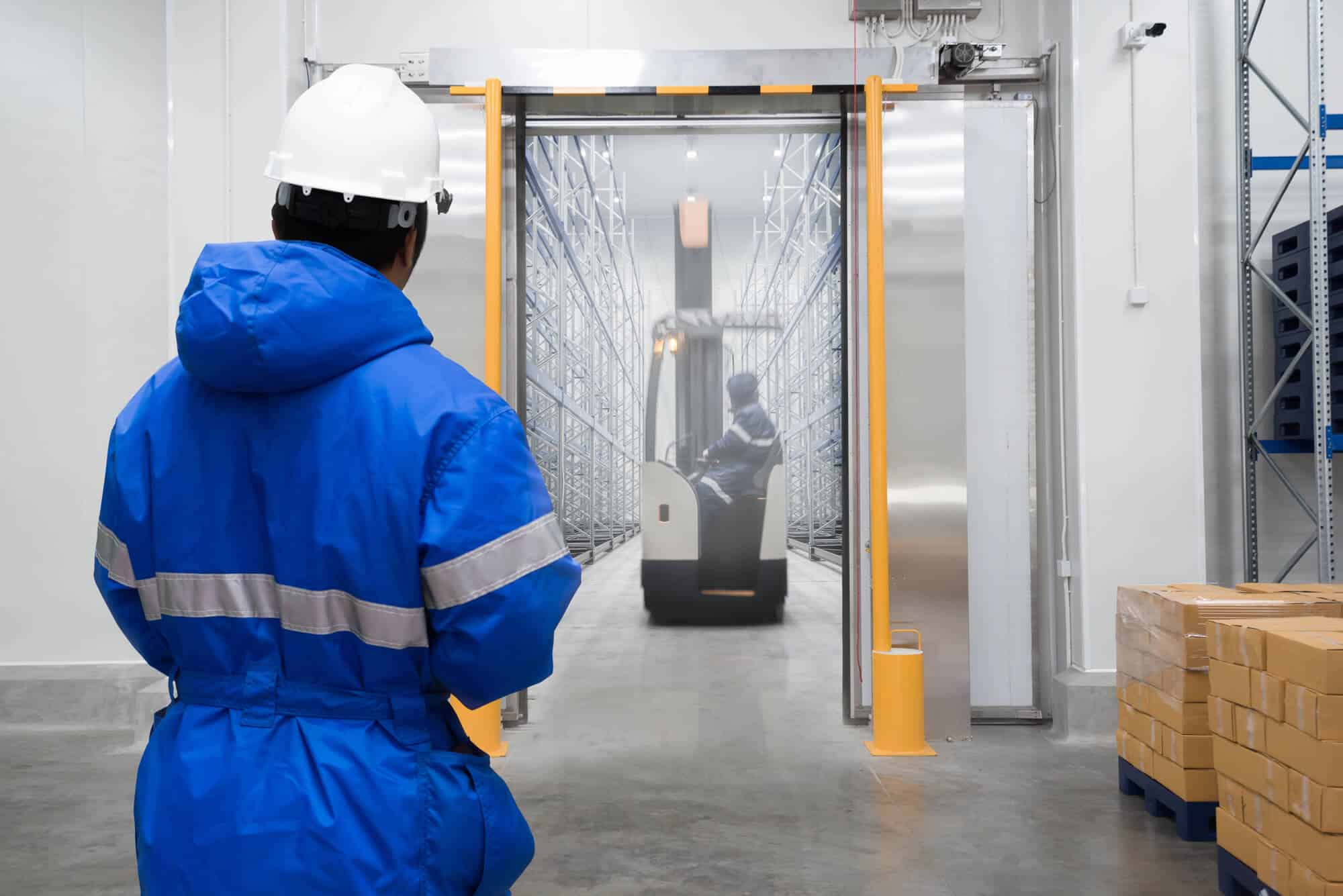 A worker in a blue jacket and white helmet watches a forklift transporting goods critical for cold chain food safety through an industrial warehouse doorway, with boxes stacked on the right.