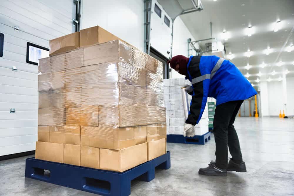 Worker in a blue jacket handling boxes inside a cold storage warehouse, organized to meet cold storage safety standards; large pallet of wrapped cardboard boxes in the foreground.
