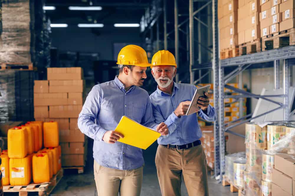 Two men in hard hats and blue shirts review documents in a warehouse, surrounded by shelves of boxes and containers, ensuring a seamless logistics solution.