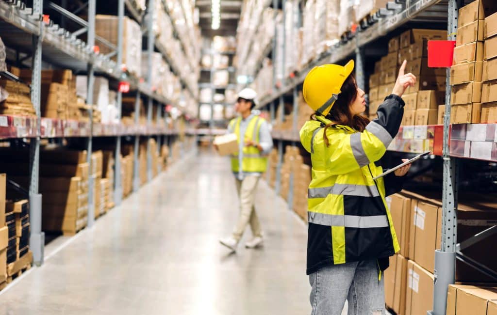 In a bustling Chicago warehouse, a worker in a high-visibility jacket and helmet points at a shelf, while another moves with precision through the cold storage area, carrying a box surrounded by stacks of cardboard cartons.