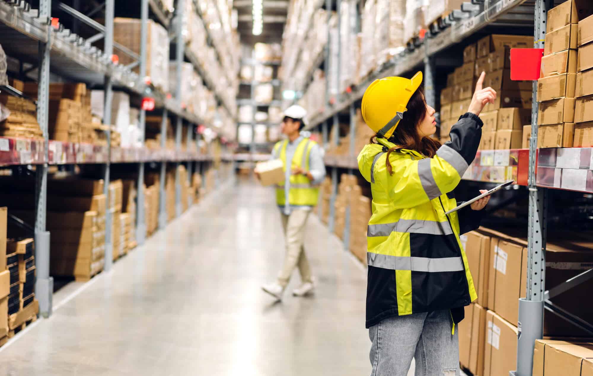 In a bustling Chicago warehouse, a worker in a high-visibility jacket and helmet points at a shelf, while another moves with precision through the cold storage area, carrying a box surrounded by stacks of cardboard cartons.