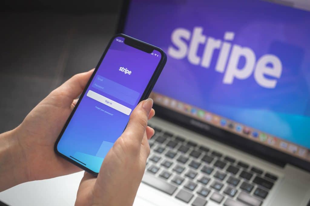 A person holds a smartphone displaying the Stripe app login screen, while in the background, a laptop shows the Stripe and Shopify logos—streamlining ecommerce and fulfillment processes.