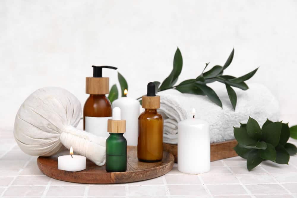 A wooden tray with amber and green glass dropper bottles, a herbal compress, lit white candles, a rolled towel, and green leaves arranged on a tiled surface—creating a soothing setting ideal for showcasing wellness products with SquareSpace Fulfillment Solutions.