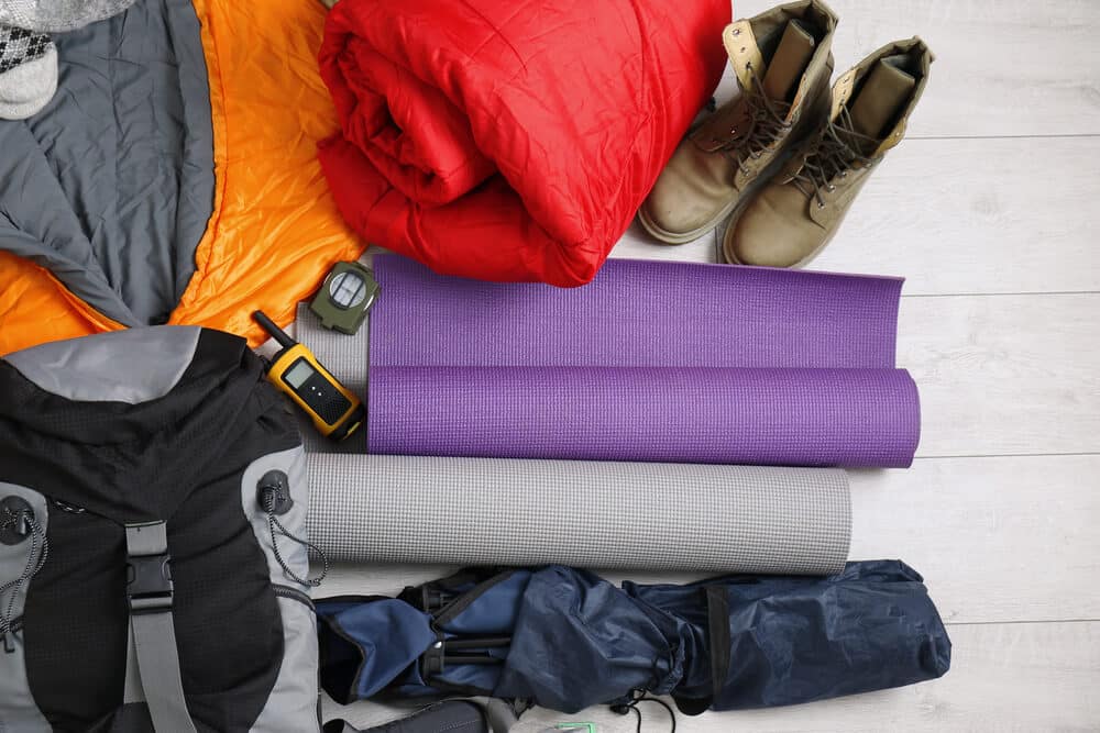 Camping gear including sleeping bags, hiking boots, a backpack, GPS device, compass, yoga mats, and a folded tent laid out on a wooden floor—organized and ready for adventure with SquareSpace Fulfillment Solutions.