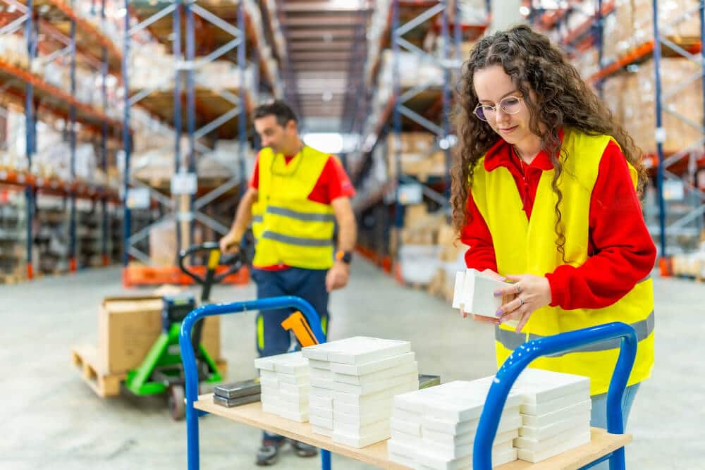 Two workers in yellow safety vests organize stacks of small boxes on a cart in a warehouse aisle, ensuring efficient Shopify Ecommerce fulfillment with shelves neatly arranged in the background.