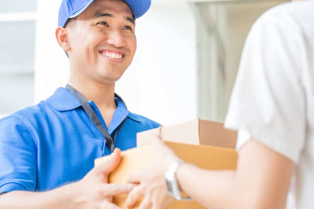 A delivery person in a blue uniform and cap hands a package to a recipient indoors, both smiling, showcasing efficient Shopify Ecommerce fulfillment in action.