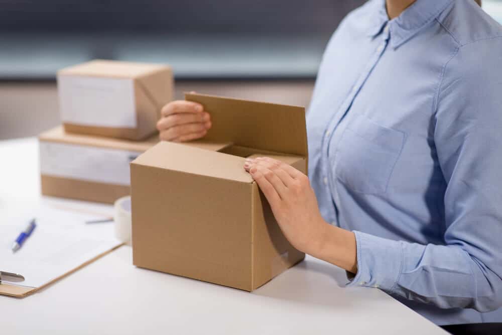 A person in a blue shirt closes a cardboard box at a table with other packages, a pen, paper, and tape visible nearby, illustrating the Shopify Ecommerce fulfillment process.