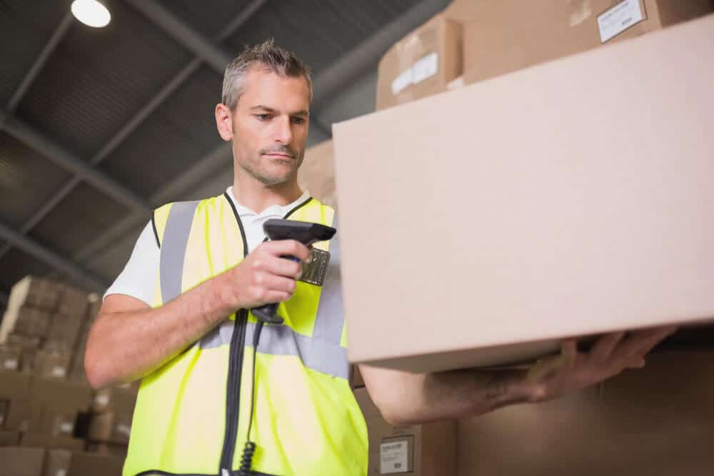 A warehouse worker in a high-visibility vest scans a barcode on a cardboard box with a handheld scanner, streamlining Shopify Ecommerce fulfillment.