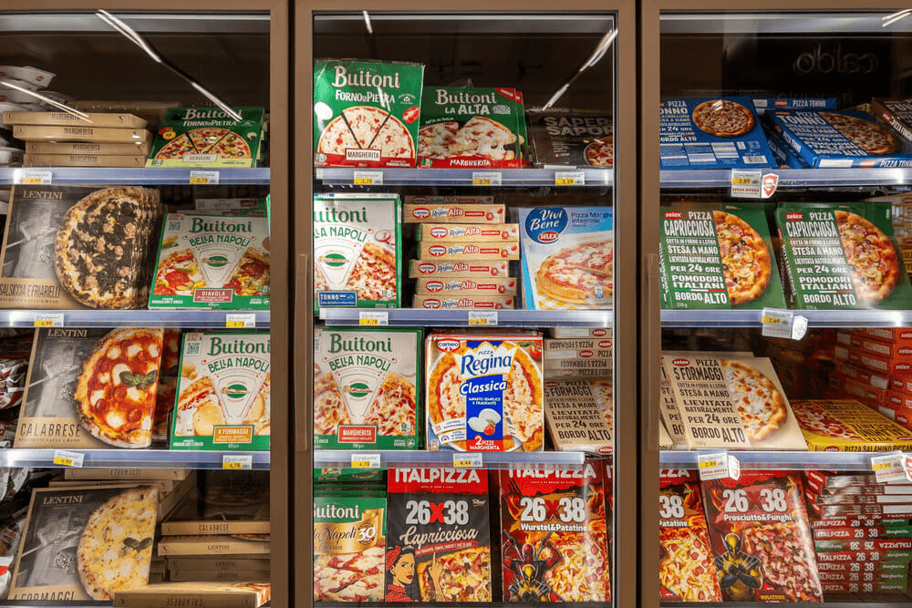 Frozen pizzas from various brands are displayed in a supermarket freezer, stacked in colorful boxes with visible labels and prices, supported by Cold Chain 3PL DHL partnership to ensure optimal freshness and reliable delivery.
