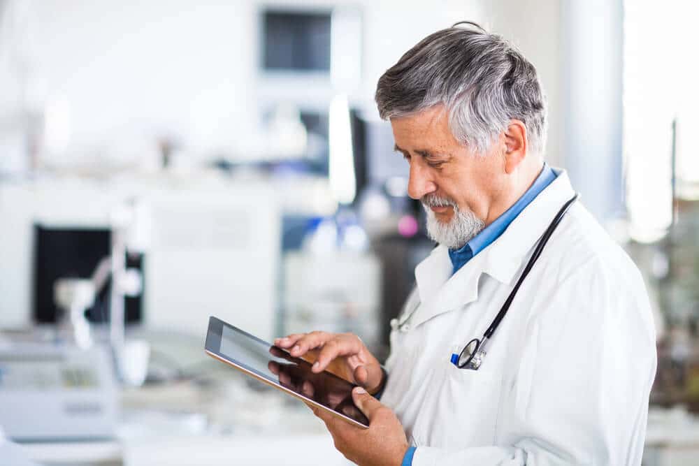 A male doctor in a white lab coat and stethoscope uses a tablet in a bright medical or laboratory setting, highlighting the benefits of Certinia supply chain integration for healthcare operations.