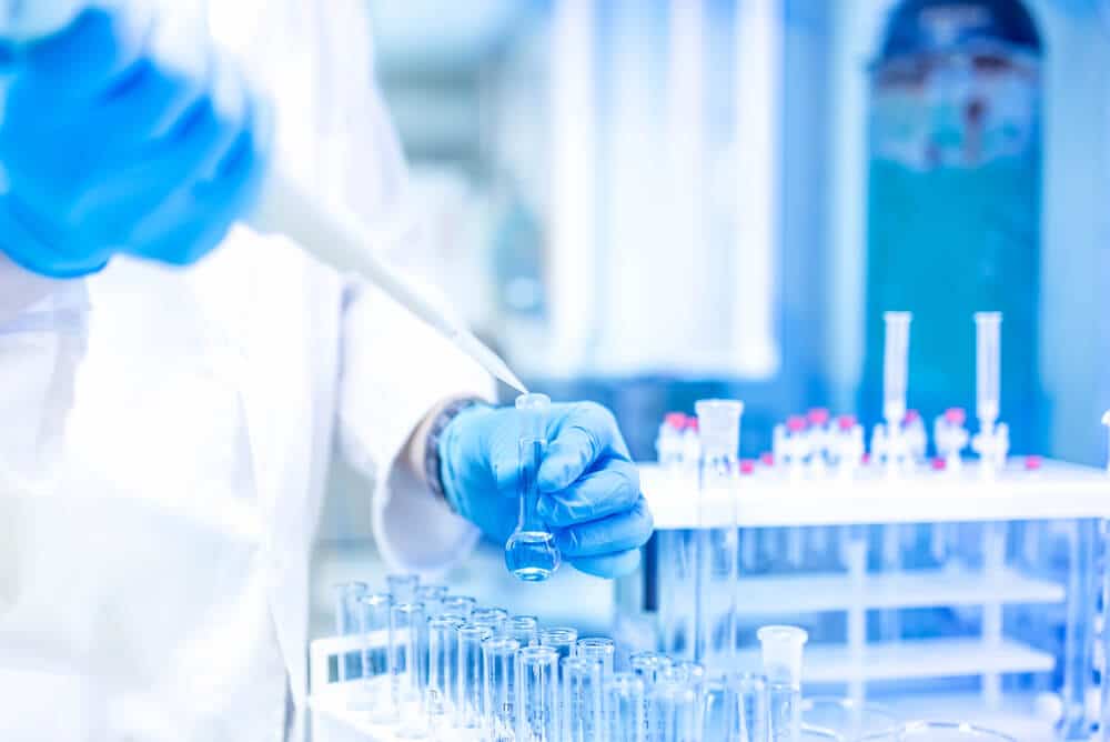A person wearing blue gloves uses a pipette to transfer liquid into a glass flask in a laboratory setting with test tubes, highlighting the importance of inventory sync with NetSuite ERP for precise lab supply management.