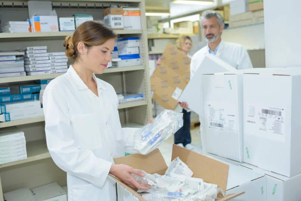 A woman in a white lab coat unpacks medical supplies from a box in a storage room, illustrating efficient DHL Fulfillment Solutions, while two others organize inventory in the background.