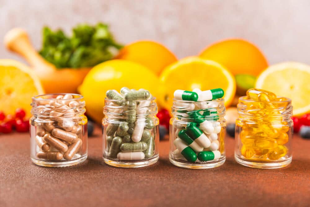 Four small glass jars filled with various capsules and softgels are lined up in front of sliced citrus fruits and leafy greens on a brown surface, symbolizing wellness products ready for international shipping with DHL and 3PL solutions.