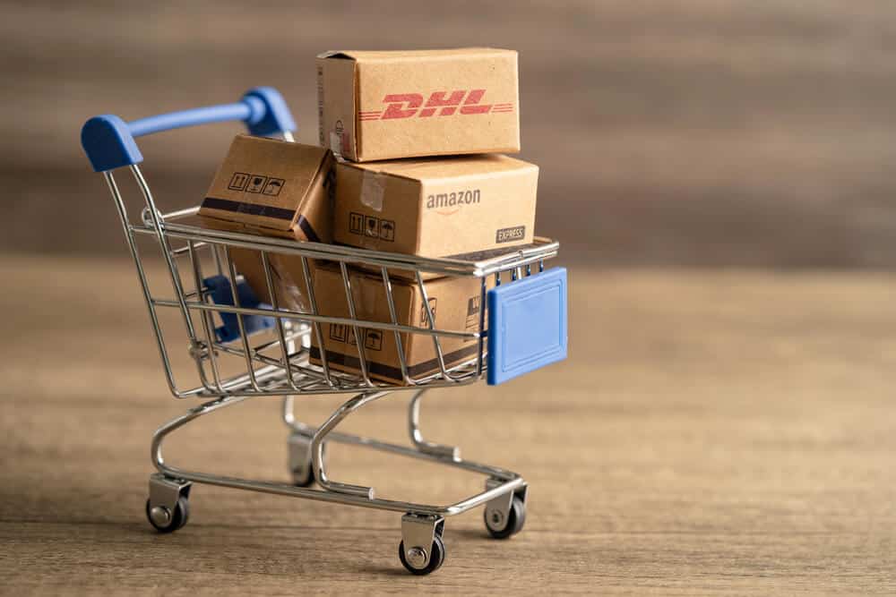 A miniature shopping cart holds small cardboard boxes labeled DHL and Amazon, showcasing DHL eCommerce logistics on a wooden surface with a blurred background.