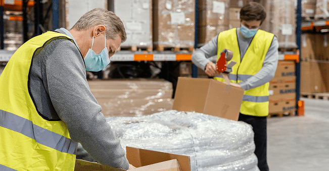Two workers wearing safety vests and face masks pack and tape cardboard boxes in a warehouse filled with shelves and wrapped pallets.
