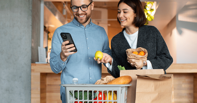 A man and woman stand indoors with groceries; he holds a phone and basket, she holds a paper bag with fruit. Both are smiling.