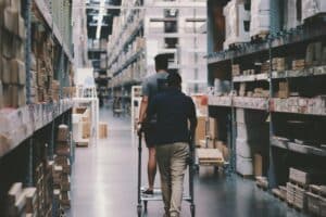 Two people move down an aisle in a warehouse, one pushing a trolley with the other riding on it; shelves are stacked with various boxes and items.
