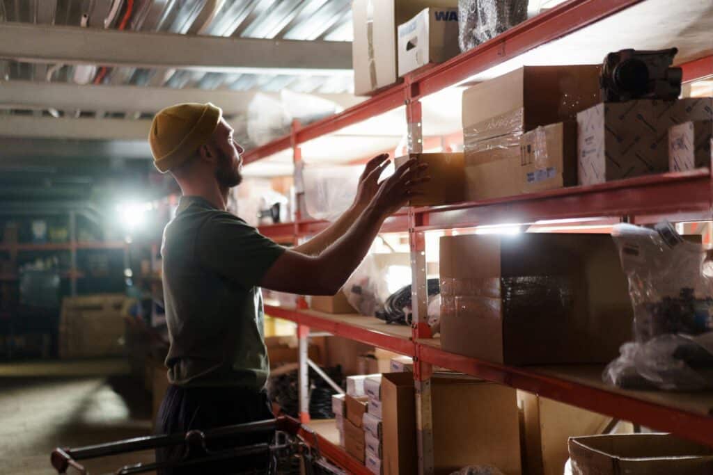 A person wearing a yellow beanie is arranging cardboard boxes on metal shelves in a warehouse with various items stored around.