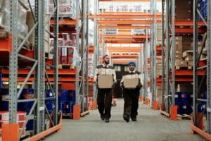 Two warehouse workers in uniforms walk down an aisle, each carrying a cardboard box, surrounded by tall shelving with stored goods.