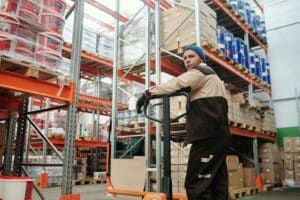 A worker in a warehouse stands next to a pallet jack, surrounded by shelves filled with boxes and supplies—an efficient scene at a third-party logistics partner handling 3PL for subscription boxes.
