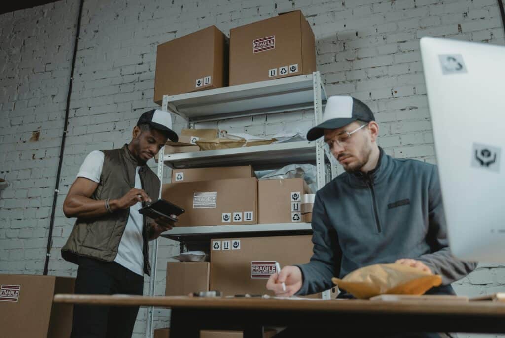 Two workers in a shipping room prepare packages for subscription box fulfillment, with "fragile" boxes stacked on shelves and a desk with a computer in the foreground.