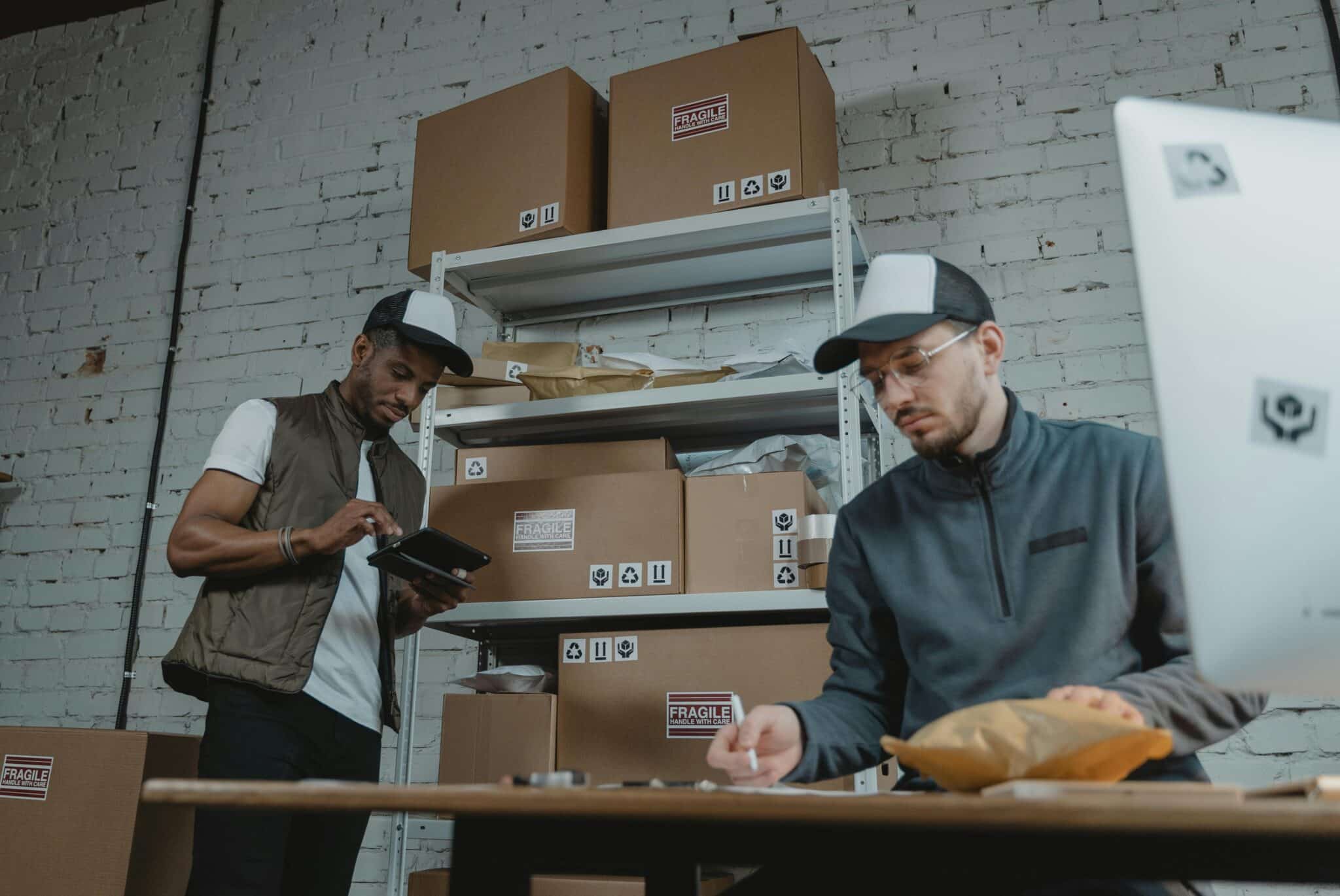 Two workers in a shipping room prepare packages for subscription box fulfillment, with "fragile" boxes stacked on shelves and a desk with a computer in the foreground.