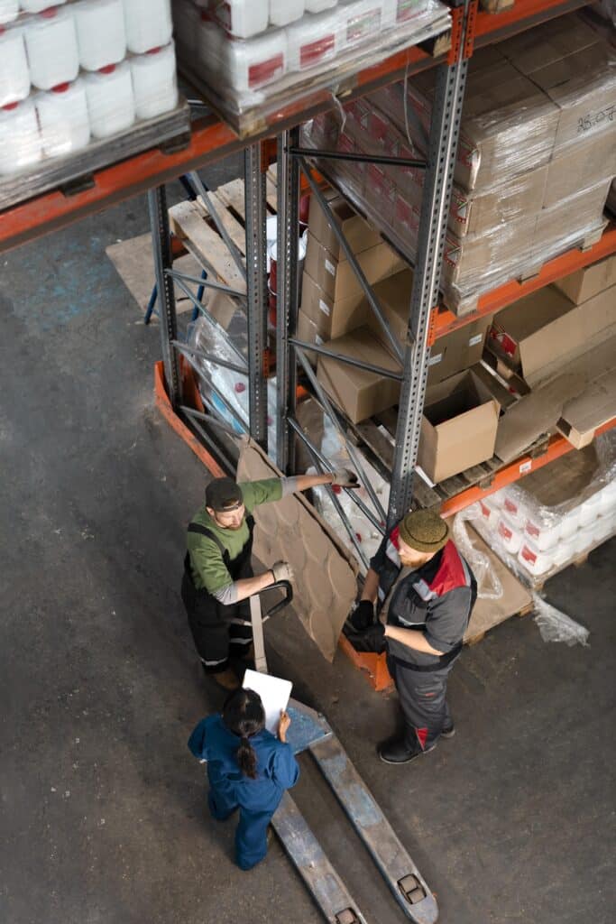 Three workers in a cold storage warehouse stand near shelves with boxes and pallets, discussing a document.